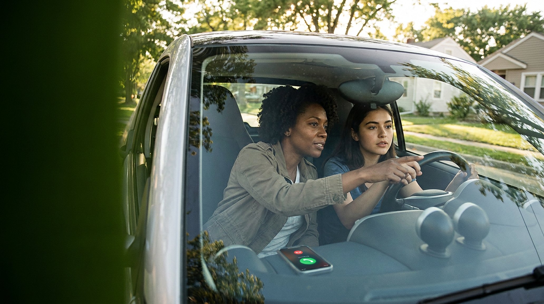 A driving instructor in the passenger seat guiding a young learner, both focused on the road; a phone lit up in the cup holder, ignored